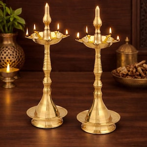 May include: Two ornate brass oil lamps with lit wicks. Each lamp has a tall, slender stem, a circular base, and a multi-tiered top with several small holders for the wicks. The lamps are on a dark wooden surface.