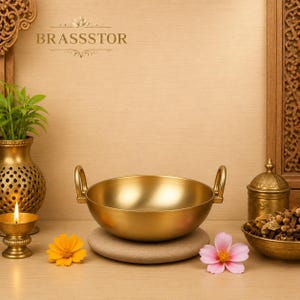 May include: A gold-coloured, round cooking pot with two handles, resting on a stone. The pot is surrounded by a candle, flowers, and other decorative items. The word "BRASSSTOR" is at the top.