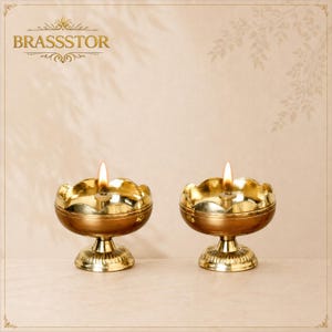 May include: Two polished brass oil lamps with scalloped edges and lit wicks. Each lamp features a wide, rounded bowl set upon a fluted pedestal base. The text BRASSTOR is visible in the upper left corner of the image, accompanied by decorative flourishes.