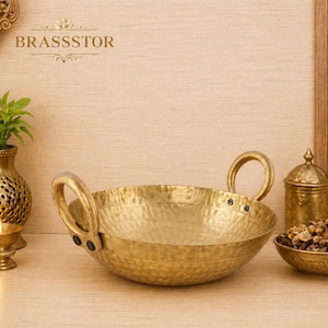 Op de afbeelding: Een gehamerde messing serveerschaal in goudkleur met twee lusvormige handvatten. De schaal heeft een getextureerd oppervlak. Een kleine messing kandelaar en een decoratieve vaas met een plant staan links. Het woord "BRASSSTOR" staat bovenaan.