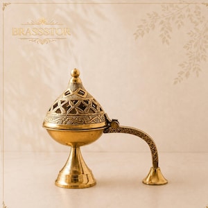 May include: A gold-colored brass incense burner featuring a dome-shaped lid with intricate cut-out geometric patterns, a smooth bowl, a pedestal base, and a curved handle with a small support stand. The text BRASSTOR is visible at the top.