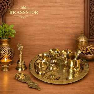 May include: A set of brass religious items, including a tray with bowls, a bell, a candle holder, and a decorative spoon. The items are arranged on a wooden surface, with the brand name "BRASSSTOR" visible.