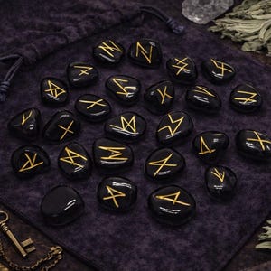 May include: A set of black rune stones with gold symbols, scattered on a dark purple cloth. A drawstring bag, a key pendant, and dried herbs are also visible. The stones are smooth and polished.