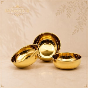 May include: Three polished gold-coloured metal bowls with a smooth, reflective finish. The bowls are arranged in a cluster, with one tilted forward and two positioned behind it. The text BRASSTOR is visible in the upper left corner of the image.