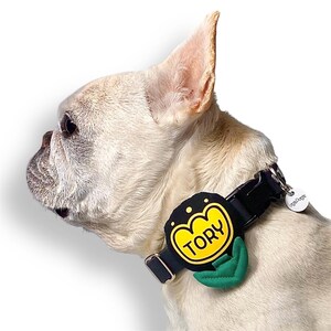 May include: A black dog collar with a yellow and black flower-shaped design. The collar has the name "TORY" in yellow letters. A green leaf design is below the flower. A small white tag with the text "ogDogge" is attached.