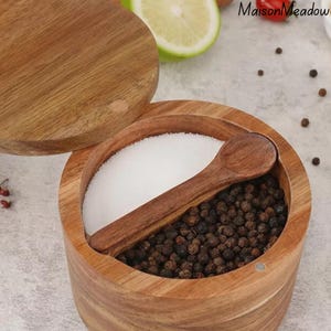 May include: A wooden salt and pepper pot with a lid and a small wooden spoon. The container is split into two compartments, one holding white salt and the other black peppercorns. The lid is partially open, revealing the contents.