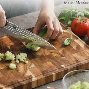 May include: A stainless steel chef's knife with a textured blade is slicing celery on a wooden cutting board. The board has a butcher block design. A tomato and basil leaves are in the background. The text "MaisonMeadow" is in the upper right corner.