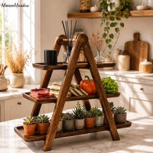 May include: A dark brown, three-tiered wooden serving stand in a ladder design. The stand holds various items, including potted succulents, sandwiches, and a black candle. The stand is on a marble countertop.