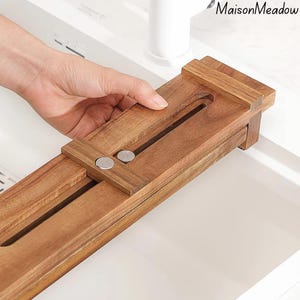 May include: A wooden kitchen sink organizer with a sliding design. The organizer is made of light brown wood and features two silver-colored magnetic closures. The text "MaisonMeadow" is in the upper right corner.