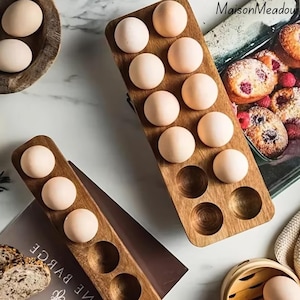 May include: Wooden egg holders with multiple egg-shaped indentations, each holding a light-coloured egg. The holders are made of a warm-toned wood. The image also shows a wooden bowl with eggs and a tray of baked goods.