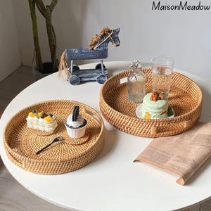 May include: Two round woven rattan trays with handles, one holding a cupcake and a slice of cake, the other holding a small cake and a glass of water. A decorative wooden horse and a glass bottle are in the background.