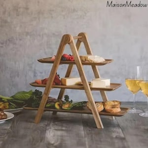 May include: A three-tiered wooden serving tray, filled with food items like cheese, berries, and bread. The tray has a triangular A-frame design. The background features a neutral-coloured wall and a wooden table. The text "MaisonMeadow" is in the upper right corner.