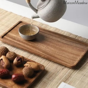 May include: A wooden serving tray with a rectangular shape. A small ceramic bowl is being filled with tea from a speckled teapot. Various nuts and dried fruits are arranged on a smaller wooden tray.