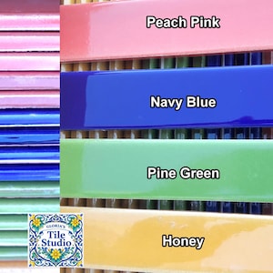 May include: A selection of glazed ceramic tiles in various colours, including peach pink, navy blue, pine green, and honey. Each tile is labelled with its colour name. The tiles are from Gloria's Tile Studio.