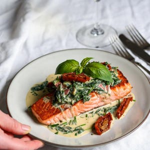 May include: A plate of salmon topped with spinach, sun-dried tomatoes, and basil. The salmon is served with a creamy sauce. A glass of white wine and silverware are in the background.