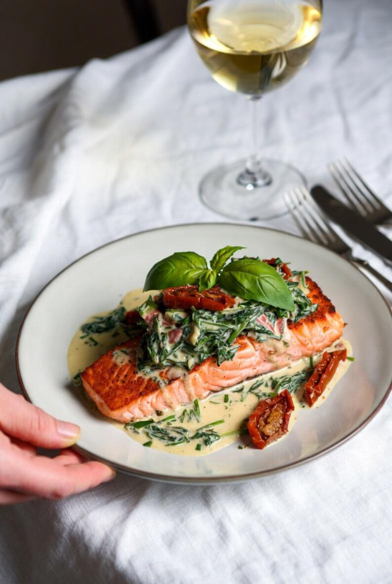 May include: A plate of salmon topped with spinach, sun-dried tomatoes, and basil. The salmon is served with a creamy sauce. A glass of white wine and silverware are in the background.