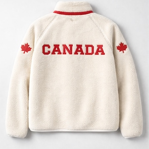 May include: Cream-colored fleece jacket with a red collar and the word "CANADA" in bold red letters across the back. Red maple leaf emblems are on each shoulder. The jacket has a high collar.
