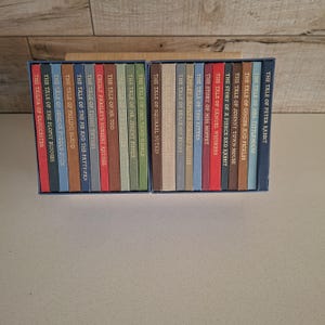 May include: A collection of twenty-three small hardback books by Beatrix Potter, arranged in two blue boxes. The spines are various colours including red, blue, green, and brown, each featuring gold-stamped titles like The Tale of Peter Rabbit.