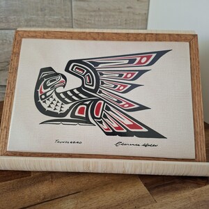 May include: A framed artwork depicting a stylized Thunderbird design. The artwork is predominantly black and red on a cream background. The frame is made of light-coloured wood. The text "Thunderbird" and a signature are visible.