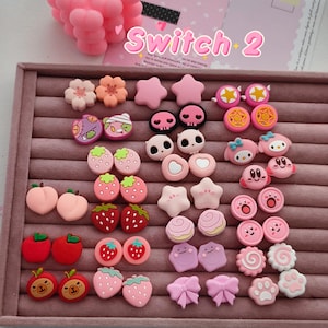 Puede incluir: Una colección de pequeñas fundas de silicona coloridas para joysticks de mandos de juego, con varios diseños que incluyen flores, estrellas, calaveras, frutas, lazos, huellas y personajes, dispuestas en una bandeja de terciopelo rosa.