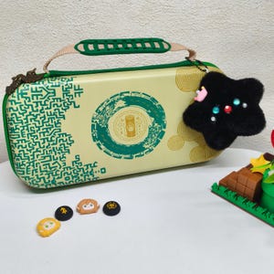 May include: A cream-colored carrying case with a green and gold design, featuring a handle and a black star-shaped charm. The case has a green zipper and a green and beige handle. Several small character-themed accessories are also visible.