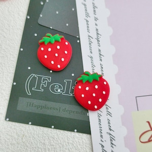 以下が含まれることがあります： 白いドットと緑色のトップが付いた、2つの赤いイチゴ型の装飾品。 「(Fel」という単語と「[Happiness] depends」というテキストが書かれた濃い緑色のカードに配置されています。背景には、筆記体のテキストが入ったピンクと白のカードが含まれています。