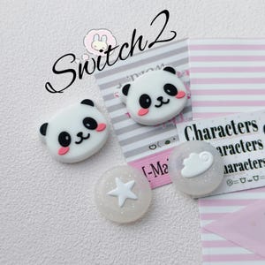 Op de afbeelding: Twee witte panda-vormige knopen met zwarte oren, ogen en roze wangen, naast twee doorschijnende ronde knopen met glitters, één met een witte ster en de andere met een witte wolk. De tekst Switch 2 is bovenaan zichtbaar.