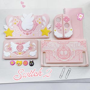 May include: A light pink gaming console accessory set featuring a dock cover, controller shells, and a console backplate with white wing motifs, yellow stars, and the text "DREAMCATCHER". Includes four small character-themed button covers.