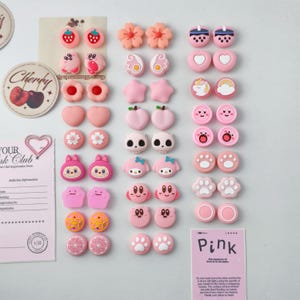 以下が含まれることがあります： イチゴ、花、ハート、漫画のキャラクターなど、さまざまな形のピンクと白の樹脂製イヤリングのコレクション。 イヤリングは白い表面に配置され、「Pink」と書かれたピンク色のカードが付いています。