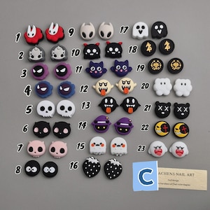 May include: A collection of 23 small, decorative nail art charms in various shapes and colors. Designs include ghosts, skulls, cats, strawberries, and other cartoon characters. The charms are arranged on a gray surface, with a business card in the bottom right corner.
