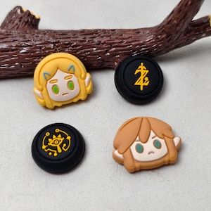 May include: Four shoe charms featuring two character faces with blonde and light brown hair, and two black circular charms with yellow symbols, including a sword emblem and a stylized eye icon, arranged on a light surface.