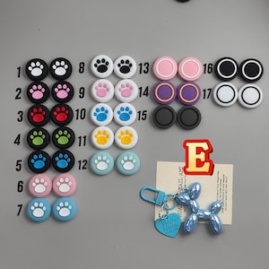 May include: A collection of colorful thumb grips for gaming controllers, featuring paw prints and solid colors. Includes a blue balloon dog keychain with a heart-shaped tag that reads "Mozzo Hiano". A red letter "E" is also present.