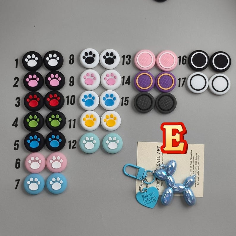 May include: A collection of colorful thumb grips for gaming controllers, featuring paw prints and solid colors. Includes a blue balloon dog keychain with a heart-shaped tag that reads "Mozzo Hiano". A red letter "E" is also present.
