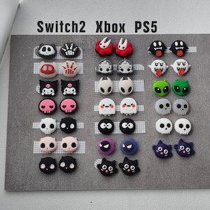 May include: A collection of small, colorful silicone charms for gaming controllers, arranged in rows under the labels Switch2, Xbox, and PS5. The charms feature various character designs, including skulls, ghosts, aliens, and animals in black, white, pink, red, green, and purple.