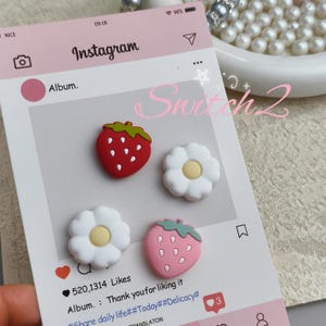 May include: Four small decorative charms shaped like strawberries and flowers, displayed on a card printed with an Instagram interface. The charms include one red strawberry, one pink strawberry, and two white flowers with yellow centers.
