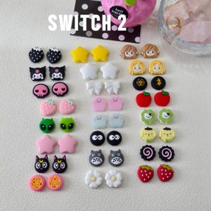 May include: A collection of colorful, decorative charms in various shapes, including stars, bows, and cartoon characters. The charms are in shades of pink, yellow, black, white, and green. The word "SWITCH 2" is printed on the top left.