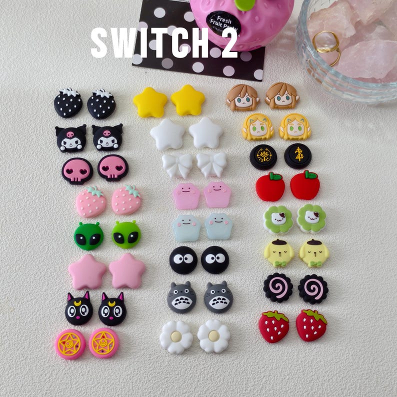 May include: A collection of colorful, decorative charms in various shapes, including stars, bows, and cartoon characters. The charms are in shades of pink, yellow, black, white, and green. The word "SWITCH 2" is printed on the top left.