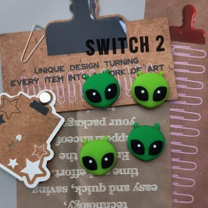 May include: Four green alien-shaped magnets with black eyes and small antennae are arranged on a brown paper background. The text "SWITCH 2" is visible, along with other text. A silver paperclip and a red clip are also present.