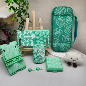 May include: A gaming console accessory set in mint green and white featuring a leaf pattern. The set includes a hard carrying case, a console dock cover, a controller shell, a game card case, a foldable stand, and four thumb grip caps.