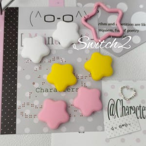 Può includere: Una collezione di ciondoli a forma di stella bianchi, gialli e rosa, accompagnati da un moschettone rosa a forma di stella e un piccolo anello argentato a forma di cuore. Gli oggetti sono disposti su carta stampata con testo e motivi a pois.