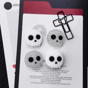 May include: Four small skull-shaped charms arranged on a white card with printed text. Two skulls are solid white, and two are translucent with silver glitter. A black metal cross-shaped paperclip is attached to the top right of the card.
