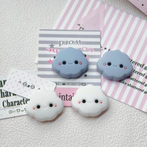 May include: Four small, cloud-shaped charms with smiling faces, two in light blue and two in white. Each cloud features two black circular eyes, a small curved mouth, and two pink dots on the cheeks. The charms are arranged on patterned paper cards.