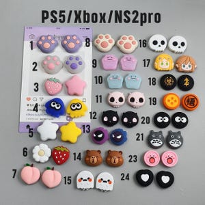 May include: A collection of silicone thumbstick covers for PS5, Xbox, and NS2pro controllers, featuring various designs including cat paws, skulls, fruits, stars, and cartoon characters, organized in a numbered grid from 1 to 24 on a gray background.