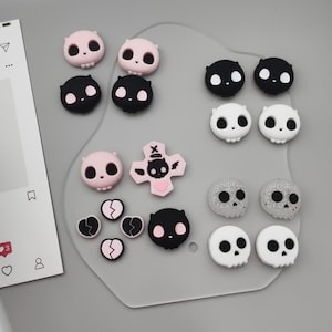 May include: A collection of various skull-shaped and heart-shaped embellishments in pink, black, white, and silver glitter. The embellishments are arranged on a gray surface, with a social media interface visible in the corner.