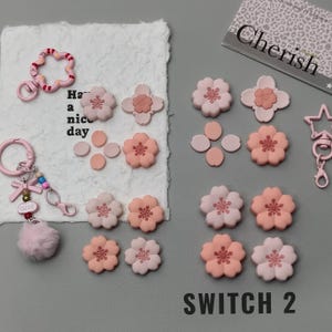 Puede incluir: Una colección de adornos y llaveros en forma de flor rosa y melocotón. Los llaveros incluyen un pompón rosa, un lazo y la palabra "cute". El texto "Have a nice day" es visible en un papel blanco.