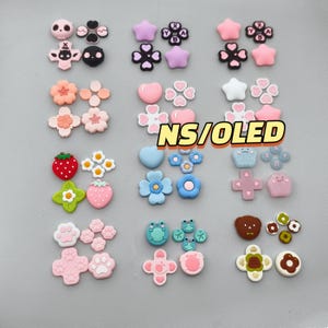 May include: A collection of colorful silicone joystick caps for gaming controllers, featuring various shapes like stars, flowers, hearts, strawberries, and animal faces, with some caps displaying the letters X, Y, A, and B, and the text NS/OLED.