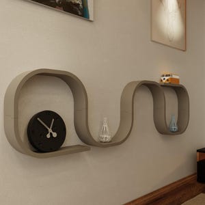 May include: A modern, taupe-colored wall shelf with a unique, flowing design. The shelf holds a black clock, decorative vases, and a small toy bus. The design is minimalist and adds a contemporary touch to any room.