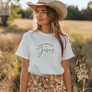 Könnte beinhalten: Ein weißes kurzärmeliges T-Shirt mit braunem Text, der "THE WAY - THE TRUTH - THE LIFE" über dem Wort "Jesus" in Schreibschrift zeigt. Das T-Shirt wird mit einem beigen Cowboyhut und einem Rock mit gelb-orangem Blumenmuster kombiniert.