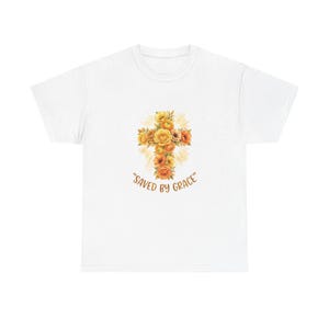 Könnte beinhalten: Ein weißes kurzärmeliges T-Shirt mit einer zentralen Grafik eines Kreuzes aus gelben und orangefarbenen Blumen vor einem hellgelben Strahlenhintergrund. Unter dem Blumenkreuz ist der Text "SAVED BY GRACE" in brauner Schrift aufgedruckt.