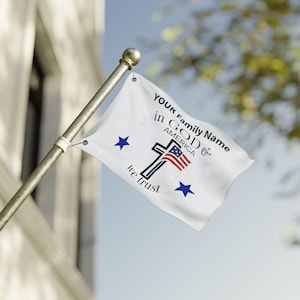 Op de afbeelding: Een witte vlag met de tekst "YOUR Family Name in GOD & AMERICA" en een kruis met een Amerikaans vlagontwerp. De vlag heeft ook twee blauwe sterren en de woorden "we trust". De vlag is bevestigd aan een zilveren paal.
